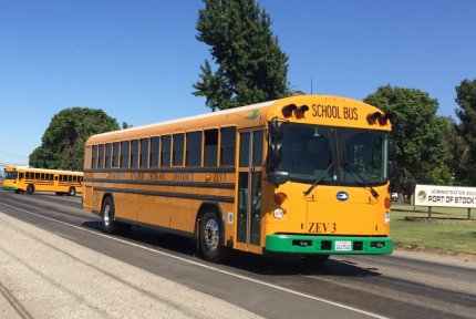 A yellow electric school bus drives down a road, as another pulls out onto the same road behind it. A sign beside the road reads: "Administration Building: Port of Stockton".